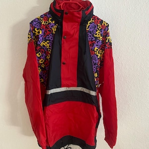 Peut inclure: Anorak vintage rouge, noir et à motifs. La veste a un col montant, un panneau central noir et un motif géométrique jaune, violet et noir sur les épaules. Une bande réfléchissante argentée traverse la poitrine.