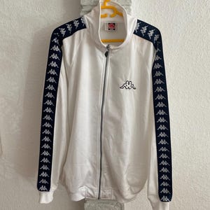 Puede incluir: Chaqueta deportiva blanca con detalles en azul marino y cremallera completa. La chaqueta presenta el logotipo de Kappa en el pecho y en las mangas. La chaqueta está colgada en una percha amarilla.