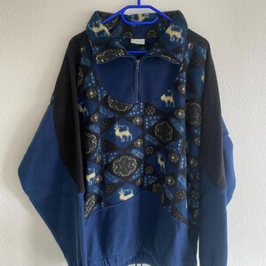Könnte beinhalten: Blauer Fleece-Pullover mit einem Muster aus Hirschen und geometrischen Formen. Der Pullover hat einen halben Reißverschluss, schwarze Ärmel und einen Kordelzug am Saum. Er hängt an einem blauen Kleiderbügel. Ca. 70 cm lang.