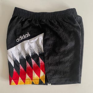 Op de afbeelding: Zwarte sportbroek met een geometrisch ontwerp en het Adidas-logo. De shorts hebben een wit, rood en geel patroon aan de linkerkant. De shorts zijn gemaakt van een textuurrijk materiaal.