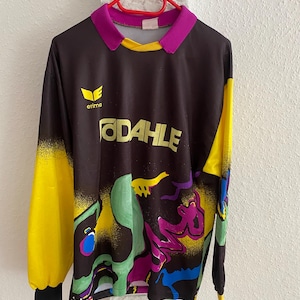 Peut inclure: Un maillot à manches longues avec une base noire, des manches jaunes et un col violet. Le maillot présente un motif abstrait coloré en vert, violet et bleu. Le mot "ODAHLE" est imprimé en jaune sur la poitrine, avec le logo "erima".