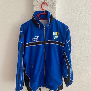 Könnte beinhalten: Blaue Jako East-Side Trainingsjacke mit schwarzen und weißen Akzenten. Die Jacke hat einen durchgehenden Reißverschluss, einen Stehkragen und die Buchstaben "CFC" auf der Brust. Die Jacke hängt an einem roten Kleiderbügel.