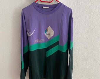 Könnte beinhalten: Ein Vintage-Sweatshirt mit langen Ärmeln in Lila, Grün und Schwarz, das an einem roten Kleiderbügel hängt. Das Sweatshirt hat das Adidas-Logo und ein geometrisches Design auf der Vorderseite. Die Ärmel sind lang und die Manschetten sind schwarz.
