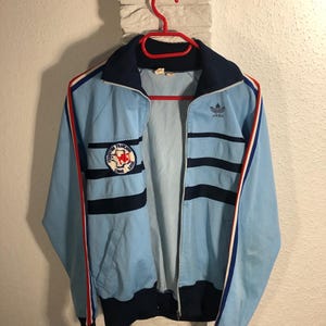 May include: A light blue vintage Adidas tracksuit jacket with red, white and blue stripes. The jacket has a zip closure and a logo patch on the left chest. The Adidas logo is on the right chest.