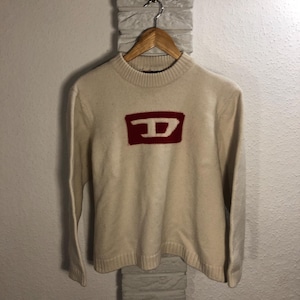 Può includere: Un maglione di lana color crema con un logo rosso e bianco con la lettera "D" sul davanti.