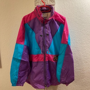 May include: A colour-blocked windbreaker jacket featuring a vibrant pattern of hot pink, bright purple, and cyan blue panels. The jacket has a high collar, a full-length front zip, long sleeves with elastic cuffs, and adjustable drawstrings.