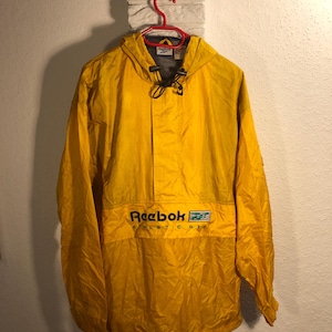 Puede incluir: Una chaqueta amarilla con capucha con un logotipo de Reebok negro y el texto "Athletic Dept." en la parte delantera. La chaqueta tiene una capucha con cordón y una cremallera.
