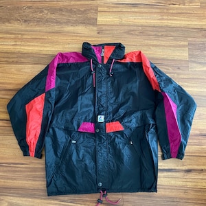 Könnte beinhalten: Eine schwarze Windjacke mit leuchtend magentafarbenen und hellorangen Farbblock-Einsätzen an den Schultern und Ärmeln. Die Jacke verfügt über einen hohen Kragen mit Kordelzug, einen Frontreißverschluss und ein kleines K-Way-Logo auf der Brust.