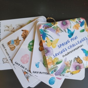 May include: A collection of white cards, bound by a metal ring, showcasing vibrant illustrations of bunnies, eggs, and butterflies. The cards display the text "Spring Mini Savings Challenges" and "Plan&Prosperbyali".