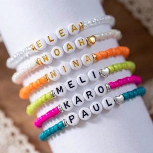 Könnte beinhalten: Eine Reihe farbenfroher Armbänder mit weißen Buchstabenperlen, die Namen buchstabieren. Die Armbänder sind in verschiedenen Farben erhältlich, darunter Silber, Orange, Grün, Pink und Türkis. Jedes Armband hat einen anderen Namen.