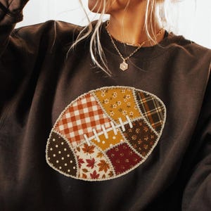 May include: A dark brown long-sleeve sweatshirt featuring a central football-shaped graphic composed of various fabric patches in autumn colors, including plaid, gingham, floral, and leaf patterns, with white stitching details on the football.