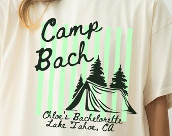 May include: An ivory short-sleeve t-shirt featuring a graphic with the text "Camp Bach" above a tent and pine tree illustration. Below the tent, the text reads "Chloe's Bachelorette Lake Tahoe, CA" over vertical light green stripes.