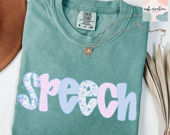 May include: A teal Comfort Colors t-shirt with the word "Speech" in a playful font. The letters are decorated with floral, gingham, and striped patterns in pink and blue. A gold necklace with a pendant rests on the shirt.