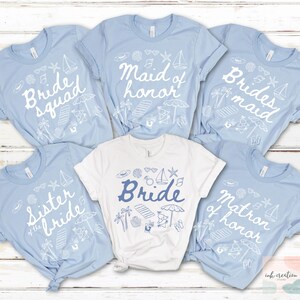 May include: A collection of light blue and white t-shirts with beach-themed graphics. The shirts feature phrases like "Bride", "Bride Squad", "Maid of Honor", and "Bridesmaid". The designs include sailboats, umbrellas, and seashells.