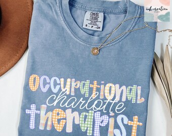 May include: A blue Comfort Colors t-shirt with the words "occupational charlotte therapist" in a colorful plaid design. A gold necklace with a pendant is draped across the shirt.