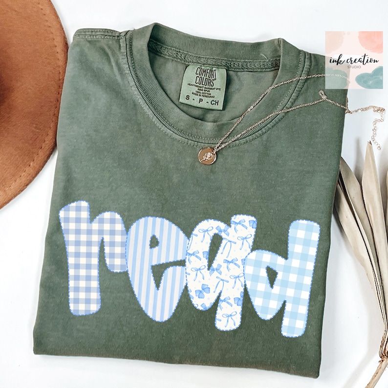 以下が含まれることがあります： オリーブグリーンのComfort Colors Tシャツ。パッチワークデザインで「read」の文字がプリントされています。文字は青と白のギンガムチェック、ストライプ、リボンのパターンです。金色のネックレスがTシャツの上に置かれています。Ink Creation Studioのロゴが見えます。