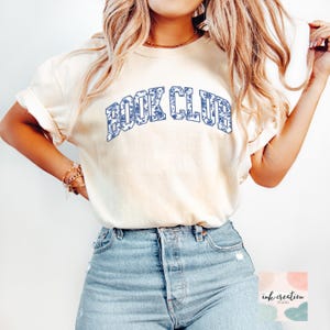 May include: A woman wearing a light beige t-shirt with the text "BOOK CLUB" in blue lettering with a floral pattern. The shirt is a relaxed fit and has a crew neck. The woman is wearing blue jeans.