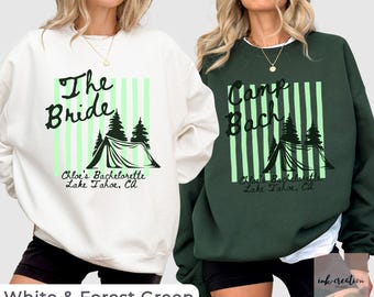 May include: A white sweatshirt and a forest green sweatshirt, each featuring a graphic of a tent and pine trees over light green vertical stripes. The white shirt reads The Bride, and the green shirt reads Camp Bach, both with Chloe's Bachelorette Lake Tahoe, CA.