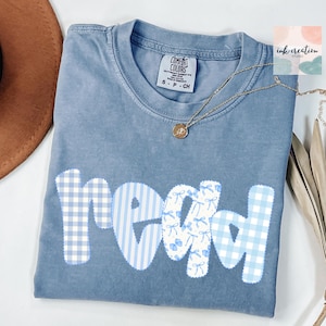 以下が含まれることがあります： パッチワークデザインで「read」の文字が入った青色のComfort Colors Tシャツ。文字は青と白のギンガム、ストライプ、リボンのパターンです。ペンダント付きのゴールドネックレスがシャツの上にあります。シャツはSサイズです。