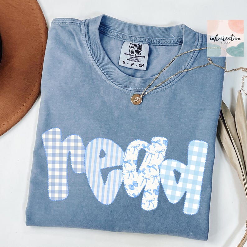 以下が含まれることがあります： パッチワークデザインで「read」の文字が入った青色のComfort Colors Tシャツ。文字は青と白のギンガム、ストライプ、リボンのパターンです。ペンダント付きのゴールドネックレスがシャツの上にあります。シャツはSサイズです。