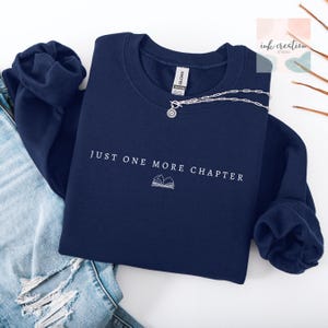May include: A navy blue sweatshirt with the text "JUST ONE MORE CHAPTER" and a book graphic in white. A silver necklace with a circular pendant is draped across the sweatshirt. The sweatshirt is folded on top of a pair of blue jeans.