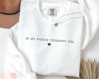 May include: White crewneck sweatshirt with the text "IN MY SPEECH THERAPIST ERA" in brown, with a brown heart below the text. A gold necklace with a pendant is draped on the sweatshirt. The sweatshirt is from Comfort Colors.