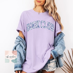 May include: A lavender t-shirt with the text "BOOK CLUB" in blue and white floral print letters. The shirt is oversized and has a relaxed fit.