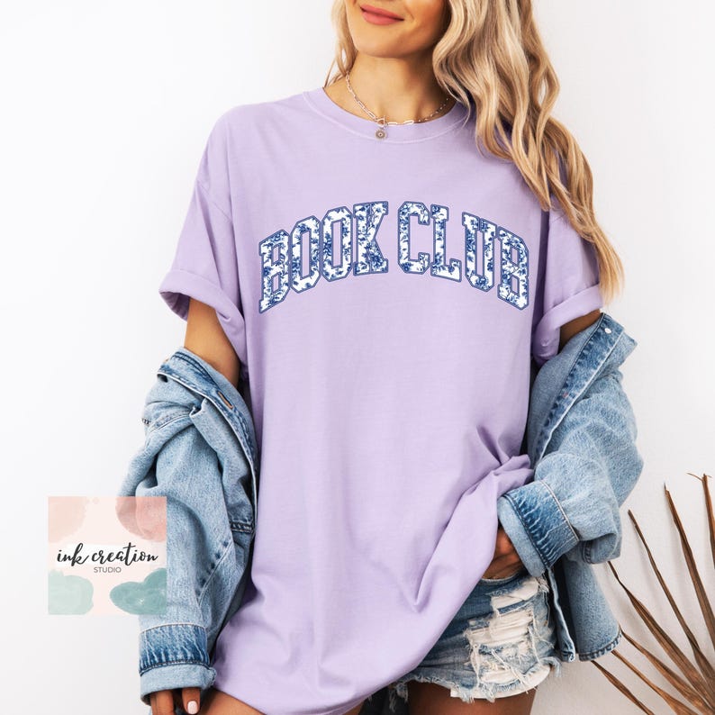 May include: A lavender t-shirt with the text "BOOK CLUB" in blue and white floral print letters. The shirt is oversized and has a relaxed fit.
