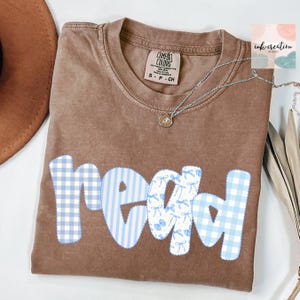 以下が含まれることがあります： 青と白の模様の文字で「read」と書かれた茶色のComfort Colors Tシャツ。「r」と「d」は青と白のギンガムチェック、「e」と「a」は青と白のストライプとリボンの模様です。ペンダント付きのシルバーネックレスがシャツの上にあります。