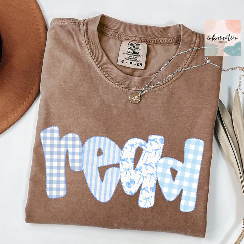 以下が含まれることがあります： 青と白の模様の文字で「read」と書かれた茶色のComfort Colors Tシャツ。「r」と「d」は青と白のギンガムチェック、「e」と「a」は青と白のストライプとリボンの模様です。ペンダント付きのシルバーネックレスがシャツの上にあります。