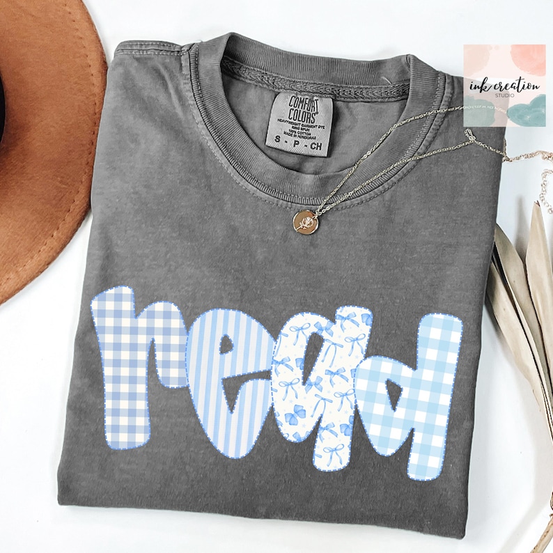 以下が含まれることがあります： 青と白の模様の生地で「read」という文字がプリントされたグレーのComfort Colors Tシャツ。「r」は青と白のギンガムチェック柄、「e」は青いストライプ、「a」は青と白のリボン柄、「d」は青と白のギンガムチェック柄。