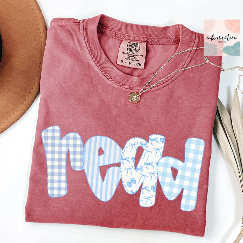以下が含まれることがあります： マルーン色のComfort Colors Tシャツ。青と白の模様の生地で「read」の文字がアップリケされています。模様にはギンガム、ストライプ、リボンが含まれています。金のペンダントネックレスがシャツの上にあります。