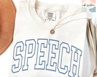 May include: Cream-colored t-shirt with the word "SPEECH" in blue block letters. A gold necklace with a pendant rests on the shirt. The shirt has a Comfort Colors tag.