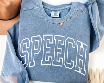 May include: A blue sweatshirt with the word "SPEECH" in white block letters. The sweatshirt has a crew neck and long sleeves. A gold necklace with a pendant is draped on the sweatshirt. A brown hat is visible in the background.
