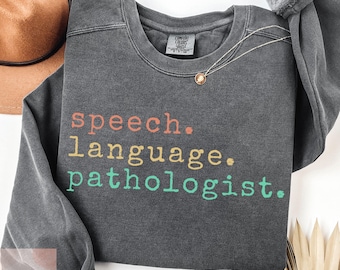 May include: A dark gray sweatshirt with the words "speech. language. pathologist." in colorful text. A gold necklace and a brown hat are visible in the background. The sweatshirt is made of a soft material.