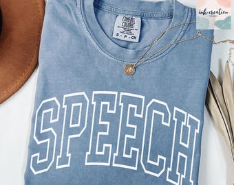 May include: A blue Comfort Colors t-shirt with the word "SPEECH" in white block letters. A gold necklace with a pendant rests on the shirt. The shirt is folded and sits next to a brown hat and dried leaves.