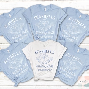 May include: Six light blue t-shirts with white text that reads "Seashells and Wedding Bells" and a graphic of seashells and wedding bells. The text also includes the bride's name, the event, and the date. One t-shirt is white with the same design.