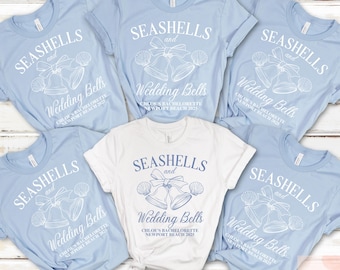 May include: Six light blue t-shirts with white text that reads "Seashells and Wedding Bells" and a graphic of seashells and wedding bells. The text also includes the bride's name, the event, and the date. One t-shirt is white with the same design.