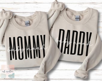 May include: Two beige crewneck sweatshirts with black text reading "MOMMY" and "DADDY". The name "baby noah" is written in a cursive font below each word. The sweatshirts are displayed on a white wooden surface.