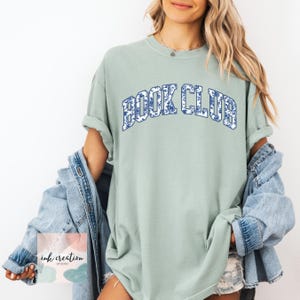 May include: A light green t-shirt with the words "BOOK CLUB" in blue letters with a white and blue floral pattern. The shirt is oversized and has a relaxed fit.