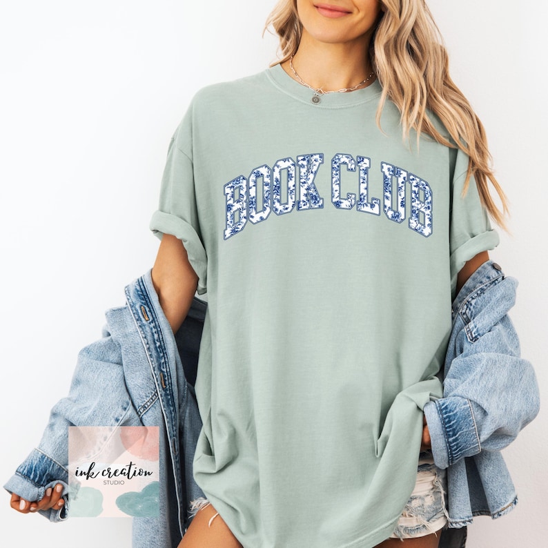 May include: A light green t-shirt with the words "BOOK CLUB" in blue letters with a white and blue floral pattern. The shirt is oversized and has a relaxed fit.