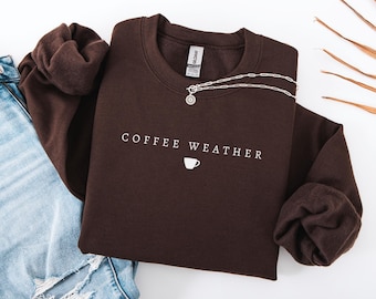 May include: A dark brown sweatshirt with the text "COFFEE WEATHER" and a coffee cup graphic in white. A silver necklace is draped across the sweatshirt. Light blue jeans are visible in the image.