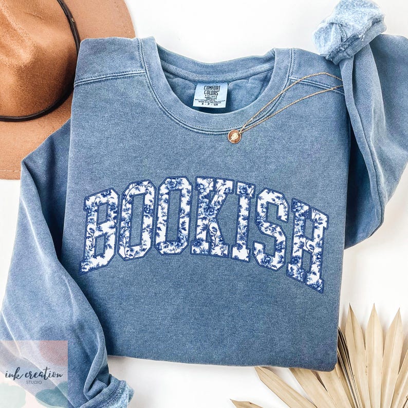 Może przedstawiać: Niebieska bluza z białym kwiatowym wzorem z napisem "BOOKISH" z przodu.