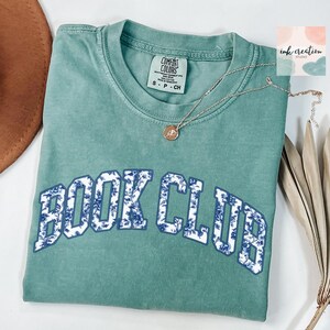 May include: A teal blue t-shirt with the words "BOOK CLUB" in white letters outlined in blue. The letters are filled with a blue and white floral pattern.