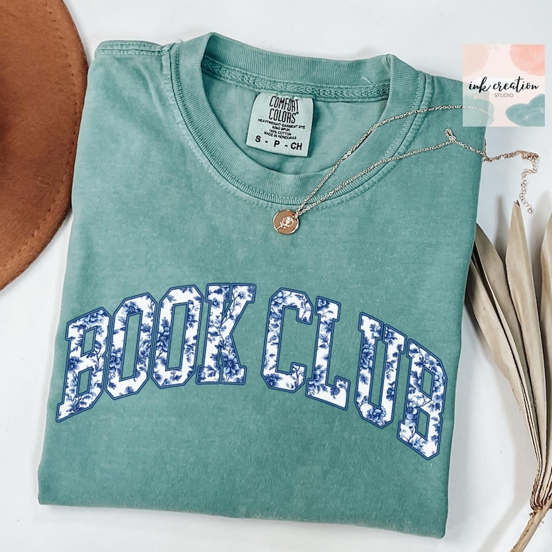 May include: A teal blue t-shirt with the words "BOOK CLUB" in white letters outlined in blue. The letters are filled with a blue and white floral pattern.