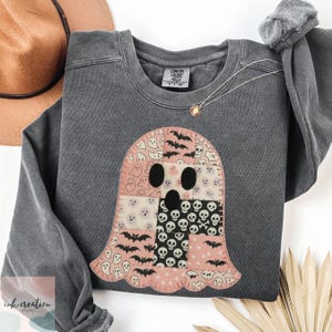 May include: Dark gray sweatshirt with a patchwork ghost design. The ghost is made of pink, white, and black fabric with bat and skull patterns. A gold necklace and a brown hat are visible.