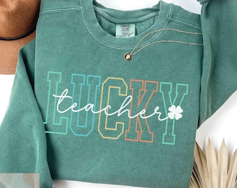 Könnte beinhalten: Grünes Sweatshirt mit dem Text "LUCKY teacher" in Regenbogenfarben mit einem vierblättrigen Kleeblatt.