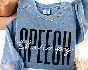 May include: A blue sweatshirt with the words "SPEECH therapy" printed in black and white. The sweatshirt has a crew neck and long sleeves. A gold necklace with a pendant is draped on the sweatshirt. A brown hat is visible in the background.