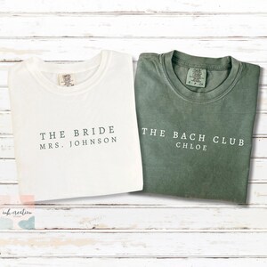 Op de afbeelding: Twee Comfort Colors T-shirts. E&eacute;n is wit met de tekst "THE BRIDE MRS. JOHNSON" in donkergroen. De andere is groen met de tekst "THE BACH CLUB CHLOE" in wit. Beide shirts liggen op een wit houten oppervlak.