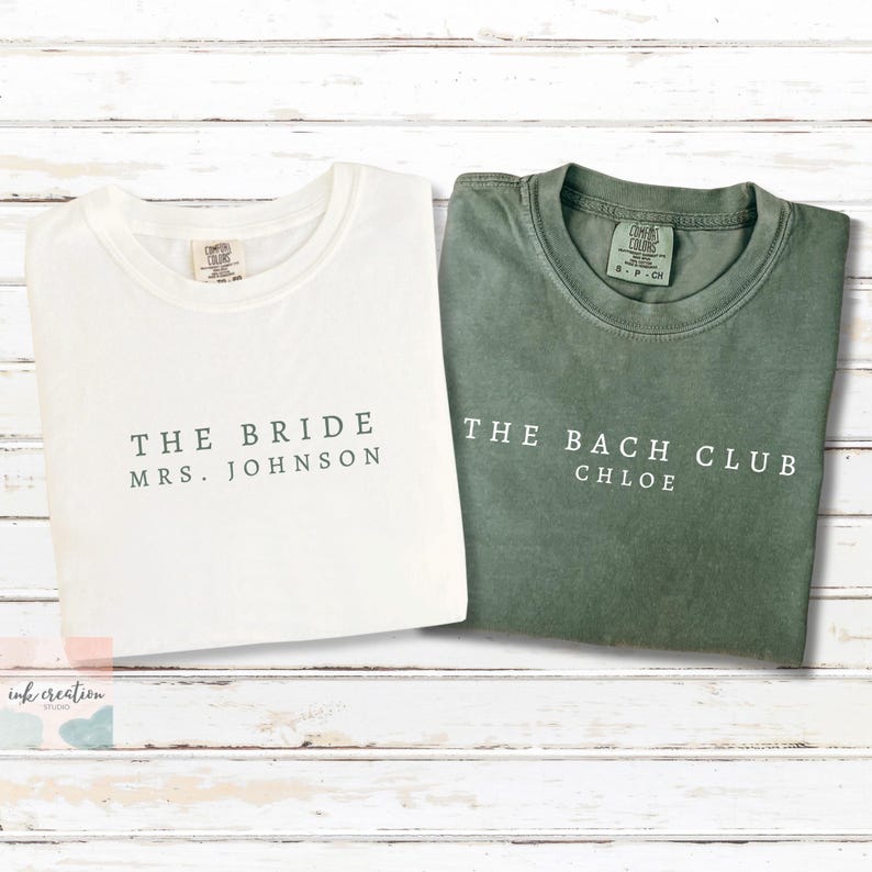 Op de afbeelding: Twee Comfort Colors T-shirts. E&eacute;n is wit met de tekst "THE BRIDE MRS. JOHNSON" in donkergroen. De andere is groen met de tekst "THE BACH CLUB CHLOE" in wit. Beide shirts liggen op een wit houten oppervlak.